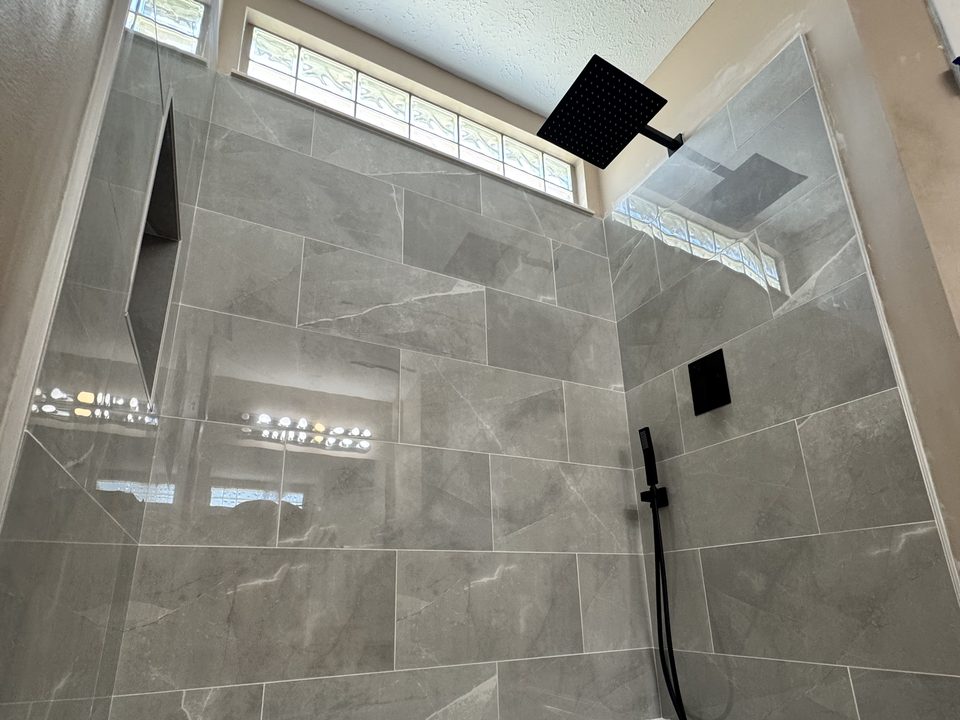 Finished gray-tile shower with glass block window, niche, and black fixtures -- bathroom floor and walls still in progress
